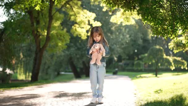 Wide shot portrait of cute Caucasian girl with toy standing in sunshine in spring summer park. Pretty happy child enjoying leisure outdoors on weekend. Childhood concept. — ストック動画
