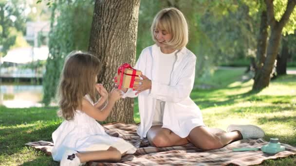 Щаслива молода жінка отримує подарунок від милої дівчинки на пікніку в сонячному парку. Портрет прекрасної кавказької матері, яка відпочиває з дочкою на відкритому повітрі у відпустці. Повільний рух. — стокове відео