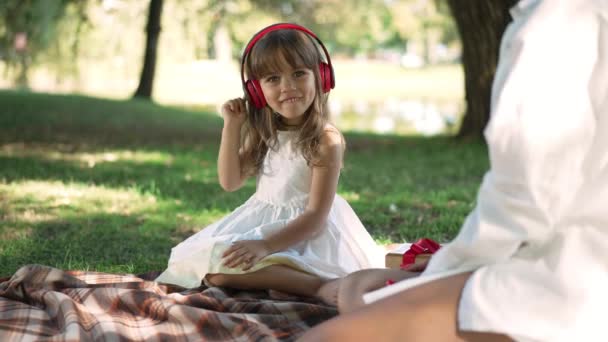 Linda niña alegre escuchando música en auriculares sentados con la mujer en el picnic de primavera y verano en el parque. Cámara en vivo zoom en la emocionada hija caucásica disfrutando de hobby al aire libre sonriendo. — Vídeo de stock
