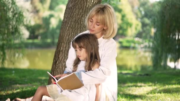 Щаслива усміхнена мати з дочкою читають книжку в сонячному парку на вулиці. Позитивна кавказька жінка і дівчинка насолоджуються відпочинком на сонці. Концепція хобі та способу життя. — стокове відео