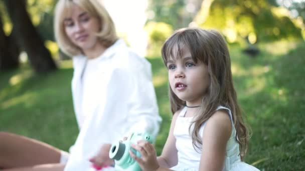 Vista lateral hábil chica curios tomar fotos con la cámara sentado con la mujer borrosa en el prado verde en el parque soleado. Encantadora hermosa hija caucásica disfrutando de hobby al aire libre descansando con la madre. — Vídeos de Stock