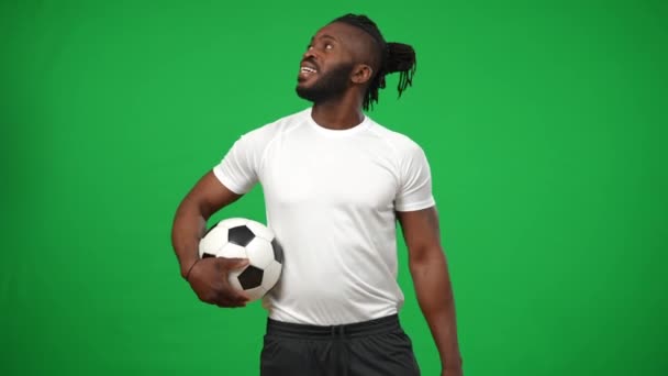 Lächelnder afroamerikanischer Sportler mit Fußballball, der aus dem Hintergrund auf eine Chromakey-Attrappe blickt und in die Kamera schaut. Porträt eines positiven selbstbewussten Mannes, der auf der grünen Leinwand posiert. — Stockvideo