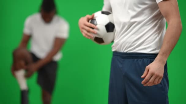 Oigenkännlig vit idrottsman studsande fotboll står till höger som afroamerikansk fotbollsspelare uppvärmning upp vid chromakey bakgrund. Självsäkra fokuserade män spelar fotboll på grön skärm. — Stockvideo