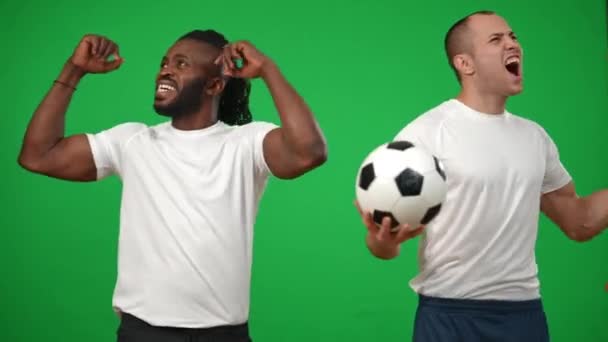 Inspirado hombres afroamericanos y caucásicos satisfechos animando a la pantalla verde mirando a su alrededor. Exitosos futbolistas seguros de sí mismos regocijándose objetivo en fondo cromakey. Concepto de deporte y éxito. — Vídeo de stock