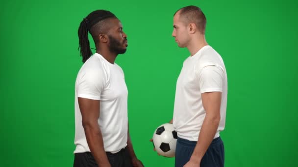 Серйозно мотивовані афро-американські та кавказькі чоловіки стояли на фоні хромакі, щоб дивитись на камеру. Портрет концентрованих спортсменів з футбольним м'ячем на зеленому екрані. — стокове відео