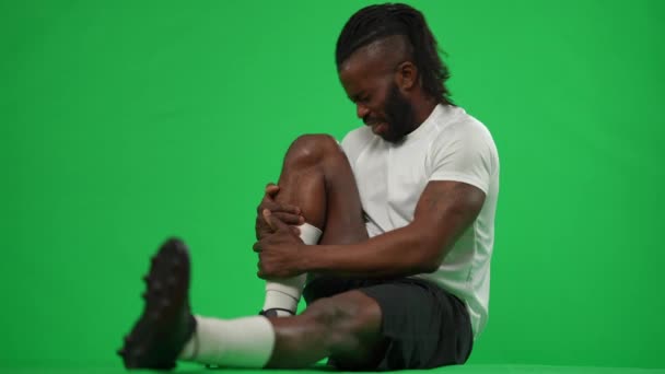 Vue de côté footballeur afro-américain assis sur l'écran vert tenant jambe blessée douloureuse. Portrait large d'un sportif stressé en arrière-plan chromatique. Concept de traumatisme sportif. — Video