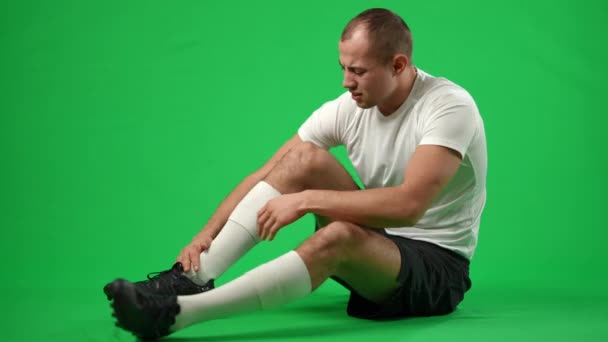 Kłopotliwy biały sportowiec siedzący na zielonym ekranie z ranną nogą. Widok z boku szeroki strzał smutny młody przystojny mężczyzna z bolesnym urazem sportowym w chromakey tle. — Wideo stockowe