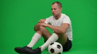 Kafkasyalı umutsuz futbolcu yeşil ekranda futbol topuyla oturup el ele tutuşarak iç çekiyor. Kromakey arka plandaki mutsuz, tatminsiz sporcunun geniş açılı portresi. Başarısızlık ve spor.