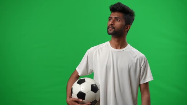 Συγκεντρωμένη Μέση Ανατολή νεαρός άνδρας με μπάλα ποδοσφαίρου κοιτάζοντας μακριά στέκεται στην πράσινη οθόνη. Νέοι επικεντρώθηκε σοβαρή αθλητής στο chromakey πρότυπο υποβάθρου. Αθλητισμός και αποφασιστικότητα. — Αρχείο Βίντεο