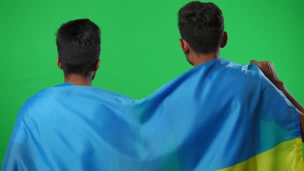 Pohled zezadu Dva muži ze Středního východu zabaleni do ukrajinské vlajky radují se z toho, že sledují fotbalový zápas na chromakey pozadí. šťastní mladí fanoušci gestikulující vítězství skákání na zelené obrazovce. — Stock video