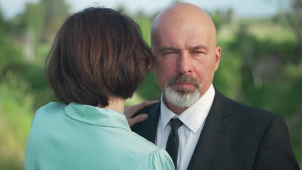Close-up portrait of handsome confident Caucasian bald man with beard and mustache looking away as woman hugging spouse outdoors. Serious elegant husband dating with wife in spring summer park. — стоковое видео