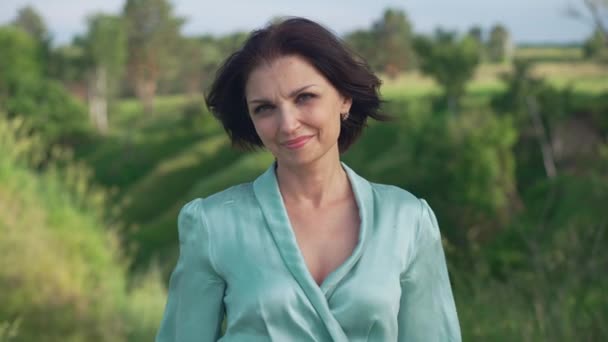 Portrait d'une jolie femme caucasienne souriante regardant la caméra debout dans le parc d'été de printemps par jour venteux. Magnifique belle dame confiante en robe élégante posant au ralenti à l'extérieur. — Video