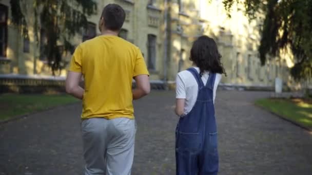 Rastreamento de companheiros de grupo caucasianos falando andando no campus universitário pela manhã. Voltar ver jovem confiante homem inteligente e mulher passeando ao ar livre discutindo estudos. Conceito de inteligência. — Vídeo de Stock