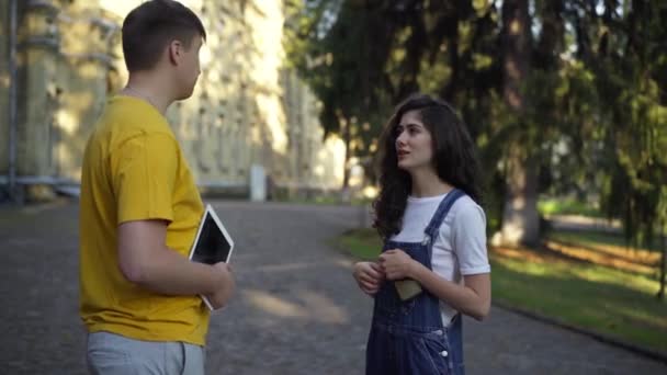 Selbstbewusste, positive junge Kaukasierin im Gespräch mit einem Mann, der draußen auf dem College-Campus steht. Glückliche intelligente Studenten, die sich morgens an der Universität unterhalten. Bildungs- und Freundschaftskonzept. — Stockvideo