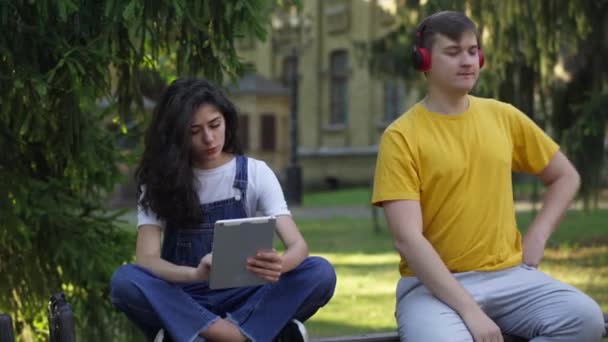 Fokuserad smal vacker kvinna studerar med tablett som avslappnad slarvig man dansar sitter på bänken på universitetsområdet. Porträtt av kaukasiska unga par utomhus på paus. Begreppet individualitet. — Stockvideo