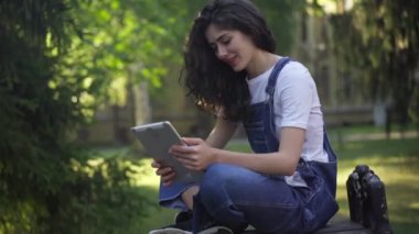 Güzel, rahat, beyaz kadın gülüyor. Tablet üzerinde sosyal medyayı seyrediyor. Üniversite bahçesinde bankta oturuyor. Üniversitenin kampüsünde sabah tatilinin tadını çıkaran kaygısız genç bir öğrenci.
