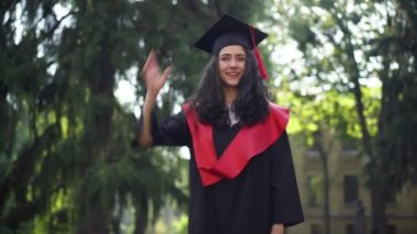 Havan topu şapkası fırlatan kendinden emin mutlu öğrenci portresi. Mezuniyet togası giymiş beyaz güzel bir kadın güneşli bir günde dışarıda dikiliyor..