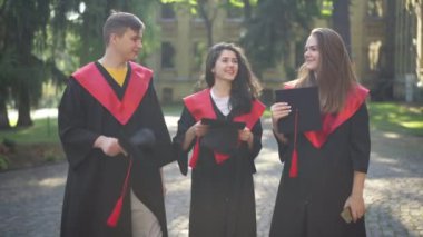 Güneşli bir sabahta havan topu şapkası atan üç mutlu öğrenci gülümsüyor. Üniversite kampüsünde açık havada poz veren mutlu beyaz erkek ve kadın portresi..