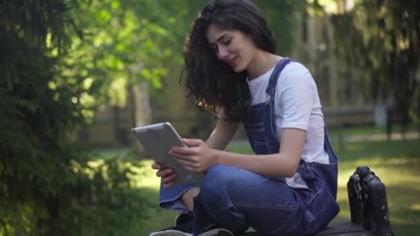 Beautiful relaxed Caucasian woman laughing watching social media on tablet sitting on bench at college yard. Positive carefree young student enjoying break in the morning outdoors at university campus — стоковое видео