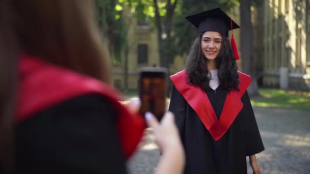 Fröhliche Absolventin wirft Mörtelmütze, während Freundin ein Foto mit dem Smartphone macht. Porträt eines glücklichen aufgeregten kaukasischen Studenten, der sich über seinen Erfolg auf dem Universitätscampus freut. — Stockvideo