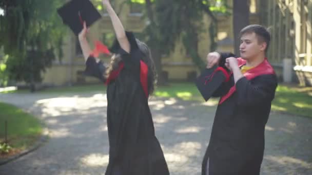 Elégedett, izgatott végzősök táncolnak a napos reggelen az egyetemi kampuszon mosolyogva. Portré boldog pozitív kaukázusi csoporttársak ballagási tógában szórakozás szabadban. Oktatás és siker. — Stock videók
