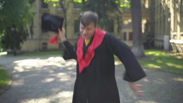 Porträtt av upphetsad ung man i examen toga dans i solsken på college campus tittar på kameran ler. Glad vit doktorand poserar ha kul utomhus på morgonen. — Stockvideo