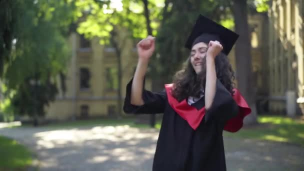 Glada unga vackra kvinna i examen toga dans på universitetsområdet på morgonen tittar på kameran. Porträtt av glada intelligenta kaukasiska doktorand poserar ha kul utomhus. — Stockvideo