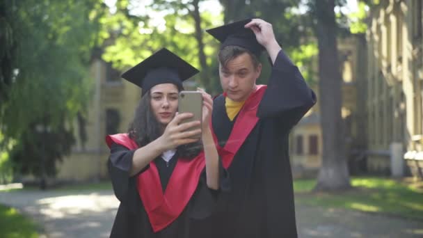 Casal satisfeito de estudantes de pós-graduação tirar selfie no smartphone em pé no campus universitário ao ar livre. Retrato de feliz namorado inteligente e namorada na graduação togas desfrutando de manhã. — Vídeo de Stock