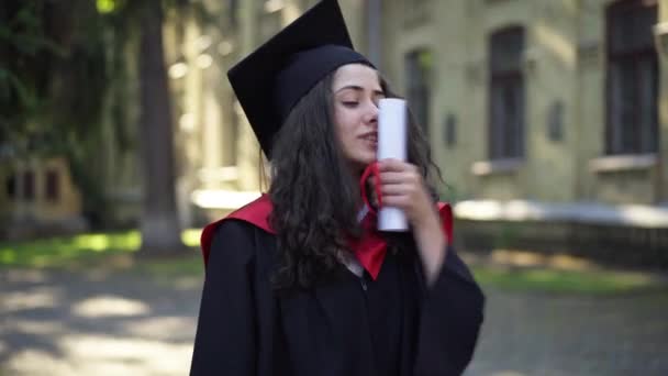 Aufgeregte intelligente schöne Frau küsst Diplom am Universitätscampus in der Früh. Porträt eines lächelnden charmanten kaukasischen Doktoranden, der seinen Abschluss im Freien genießt. — Stockvideo