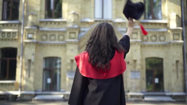 Pohled zezadu štíhlá mladá žena v maturitní tóze mávající na rozloučenou minometnou čepicí univerzitnímu kampusu. Šťastný běloch absolvent student venku na kolejích v dopoledních hodinách. — Stock video