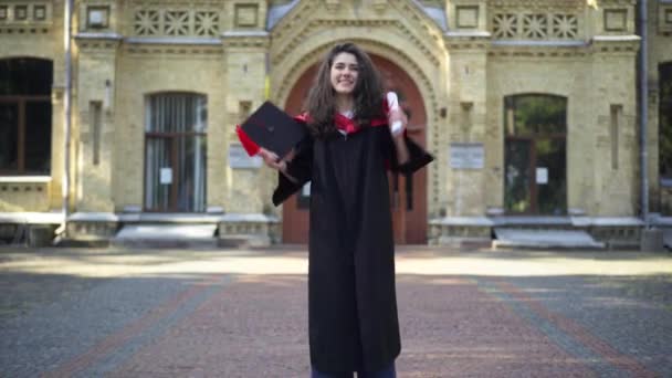 Excité satisfait belle femme dans la remise des diplômes toga saut réjouissant en regardant la caméra souriant. Portrait de heureux étudiant caucasien diplômé de l'université posant sur le campus en plein air. — Video