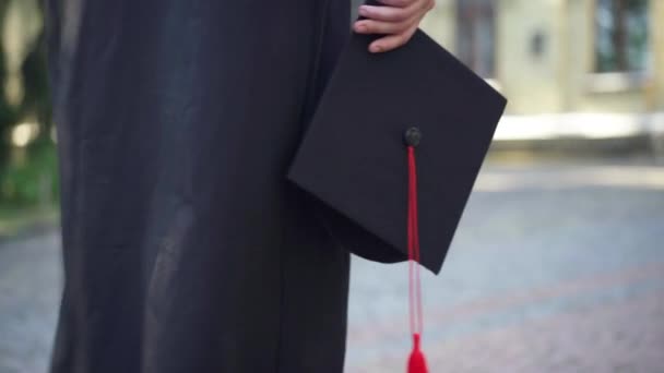 Close-up mortarboard cap in hand of unrecognizable female student walking at university campus outdoors. Young Caucasian slim woman in black graduation toga strolling at college. Tracking shot. — 비디오