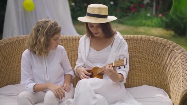 Young slim beautiful woman playing ukulele in slow motion as curios teenage girl listening song sitting on couch with sister. Caucasian talented musician enjoying leisure with sibling outdoors. — Stock Video