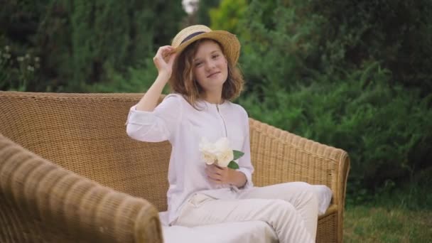 Уверенная девушка-подросток в соломенной шляпе сидит на диване под открытым небом с букетом белых роз, смотрящих на улыбающуюся камеру. Портрет счастливого симпатичного подростка, позирующего на заднем дворе в замедленной съемке. — стоковое видео