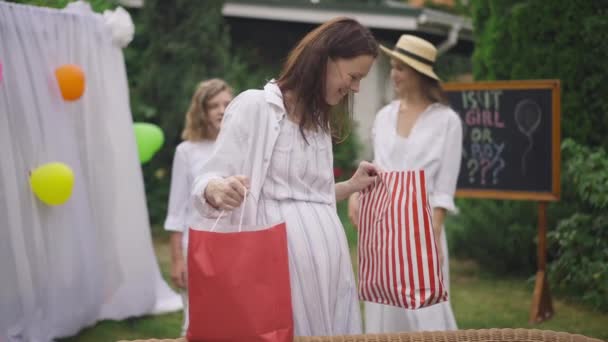 Femme enceinte caucasienne excitée se réjouissant des cadeaux debout avec des sacs à provisions sur la cour arrière comme des filles geste haut-cinq en arrière-plan. portrait de mère heureuse célébrant bébé douche. Mouvement lent. — Video