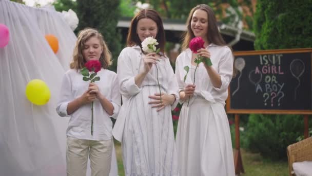 Lächelnde schöne kaukasische Frauen und Mädchen, die in Zeitlupe Rosen riechen und draußen auf dem Hinterhof stehen. Porträt von vorn: selbstbewusste schwangere Mutter und charmante Töchter feiern Babydusche. — Stockvideo