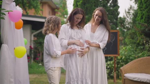 Smiling young and teenage daughters touching pregnant belly of happy mother standing on backyard. Positive Caucasian woman and girl supporting parent outdoors celebrating baby shower. — ストック動画