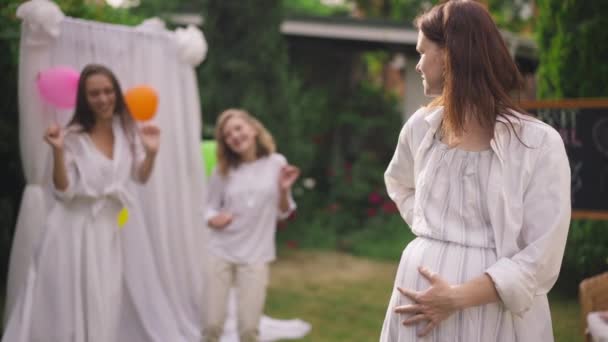 Retrato de mulher grávida caucasiana feliz ruiva acariciando barriga em câmera lenta com irmãs borradas dançando no fundo. Sorrindo satisfeito expectante celebrando festa do chuveiro do bebê com filhas. — Vídeo de Stock