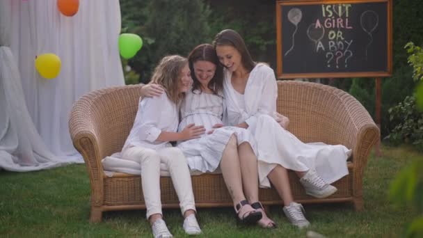 Charming happy pregnant woman and girls sitting on couch on backyard celebrating baby shower party. Wide shot smiling Caucasian teenage and young girl touching belly in slow motion. — ストック動画