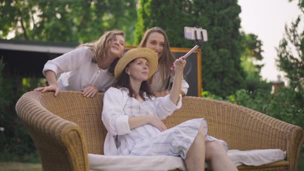 Pregnant Caucasian woman in straw hat and white dress sitting on couch taking selfie with cheerful girls grimacing in slow motion. Happy mother having fun with pretty daughters on backyard outdoors. — ストック動画