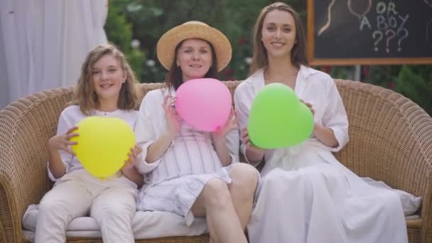 Vooraanzicht portret van vrolijke zwangere vrouw en schattige meisjes kijken naar camera gooien kleurrijke ballonnen in hartvorm lachen. Gelukkig ontspannen Kaukasische moeder en dochters vieren baby shower. — Stockvideo