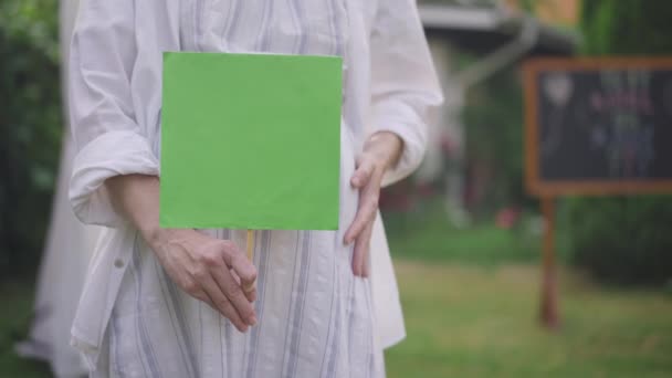 Неузнаваемая белая беременная женщина стоит на улице, держа макет зеленого знака. Уверенный ожидания на весеннем летнем дворе с шаблоном. Медленное движение. — стоковое видео
