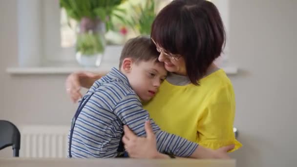 Älskade autistiska son kramar mamma sitter med förälder vid bordet inomhus. Porträtt av söt vit pojke med födelse anomali omfamna kvinna hemma. Kärlek och familjebegrepp. — Stockvideo