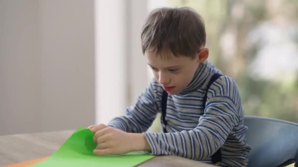 Curios autistiska kaukasiska pojke sitter vid bordet med färgglada papper ler bort. Porträtt av söta barn med psykisk störning njuter fritid hemma. Medicin och hälsa. — Stockvideo