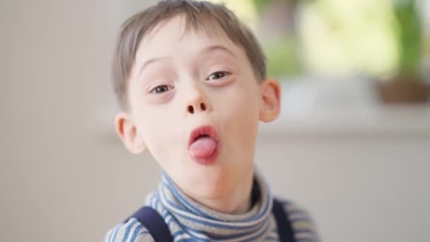 Headshot portræt af positiv autistisk dreng viser tungen ud at kigge på kameraet og griner ser væk. Close-up munter kaukasiske barn med psykisk lidelse poserende indendørs have det sjovt. – Stock-video