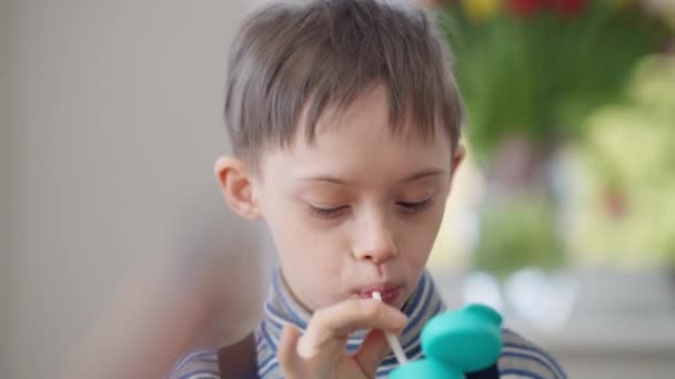 Headshot portrait of cute autistic boy drinking juice passing bottle smiling. Close-up charming Caucasian child with birth anomaly enjoying organic vitamin drink indoors at home. — стоковое видео