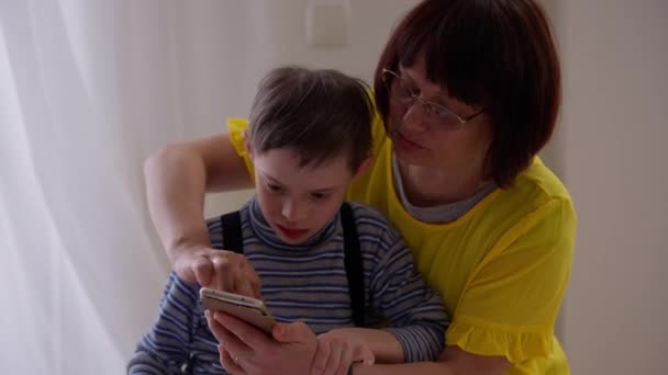 Mère caucasienne étreignant fils autiste montrant des jeux sur smartphone parler. Portrait d'une femme confiante bavardant avec un enfant souffrant de troubles mentaux à la maison à l'intérieur. Concept de jeu et d'autisme. — Video