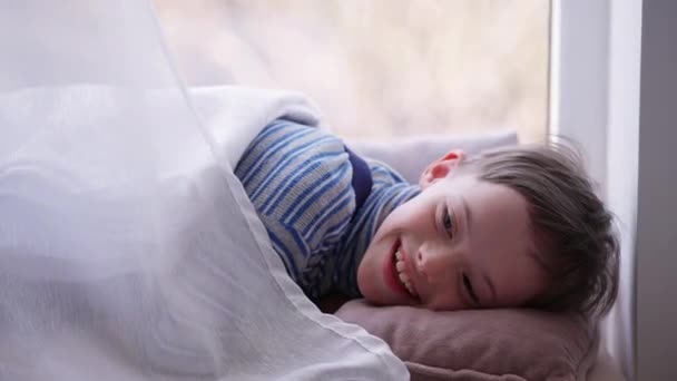 Portret van positieve schattige lachende autistische jongen liggend op vensterbank weg te kijken. Gelukkig ontspannen blank kind met geboorte anomalie psychische stoornis genieten van vrije tijd thuis binnen. Autisme. — Stockvideo