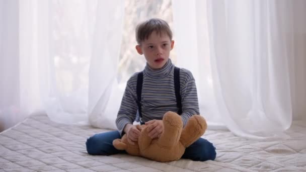 Wide shot autistic boy with teddy bear sitting on bed at home showing tongue out playing. Portrait of relaxed curios Caucasian child with birth anomaly resting indoors in bedroom. — ストック動画