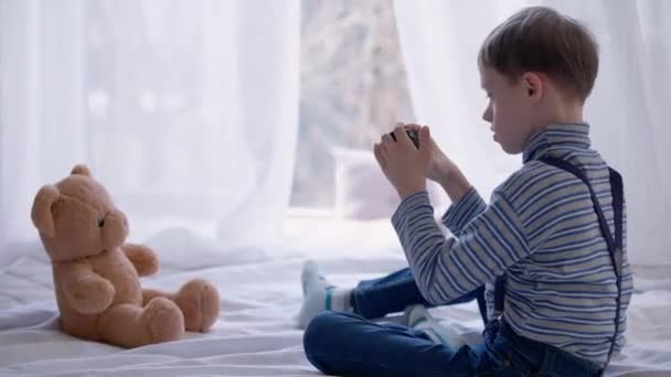 Zijaanzicht breed schot van autistische jongen het nemen van foto van teddybeer zitten met speelgoed op bed thuis. Curios geconcentreerd getalenteerd kind met psychische stoornis fotograferen genieten hobby. — Stockvideo